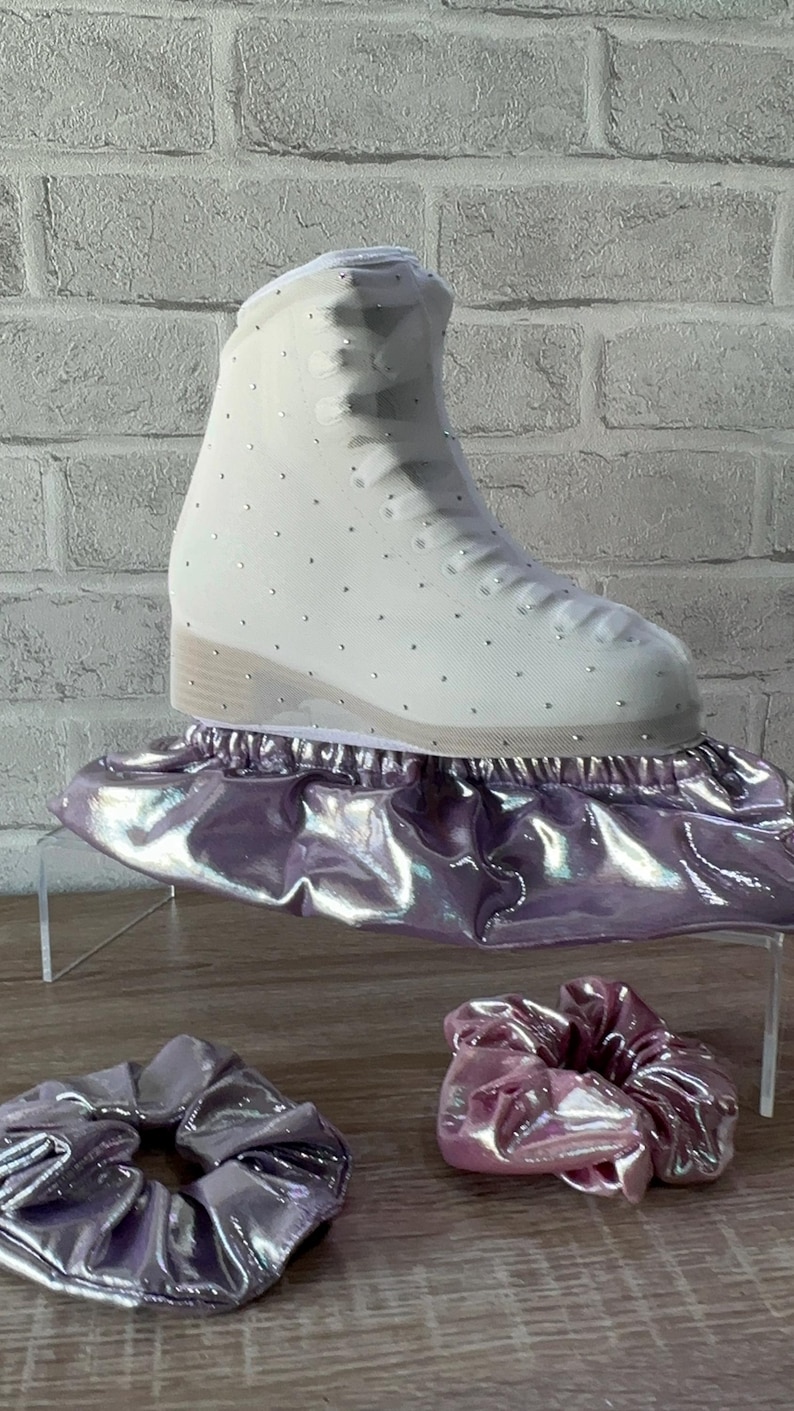May include: A white ice skate boot covered in small silver crystals, resting on a metallic lavender skate cover. Two matching metallic lavender scrunchies are positioned in front of the skate on the surface.