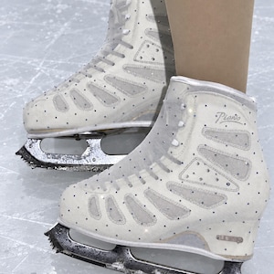 May include: A pair of white ice skates featuring mesh panels and scattered clear crystals. The boots have white laces and the word Piano printed in silver script on the side. The skates are equipped with metal blades attached to the soles.