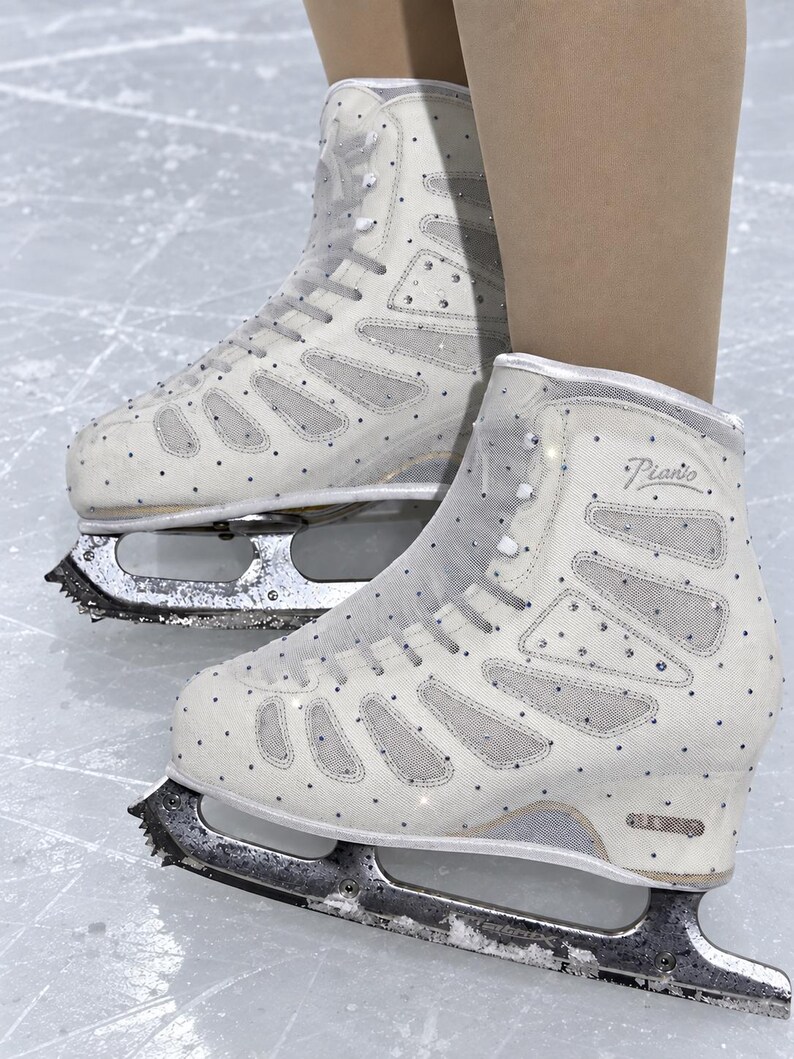 May include: A pair of white ice skates featuring mesh panels and scattered clear crystals. The boots have white laces and the word Piano printed in silver script on the side. The skates are equipped with metal blades attached to the soles.