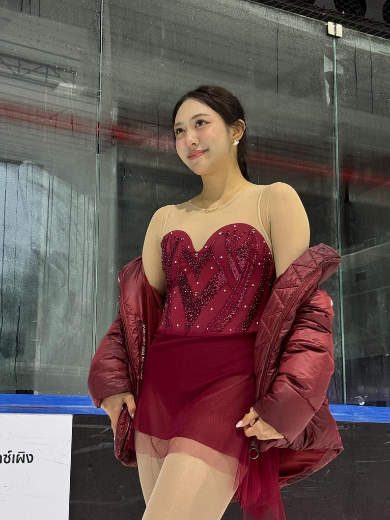 May include: A maroon figure skating dress with a sweetheart neckline, sheer nude-colored upper bodice, and intricate sequin detailing. The dress is paired with a matching maroon quilted puffer jacket draped over the shoulders.