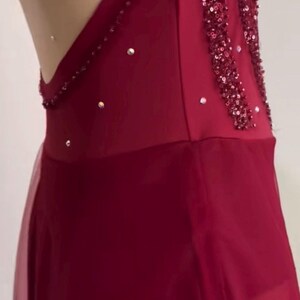 May include: A deep red sleeveless garment featuring a sheer nude-colored mesh panel along the side, embellished with vertical rows of dark red sequins and scattered clear crystals. The lower portion consists of a layered, semi-sheer red fabric skirt.