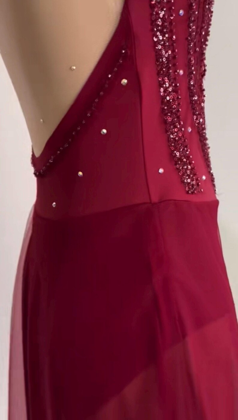 May include: A deep red sleeveless garment featuring a sheer nude-colored mesh panel along the side, embellished with vertical rows of dark red sequins and scattered clear crystals. The lower portion consists of a layered, semi-sheer red fabric skirt.