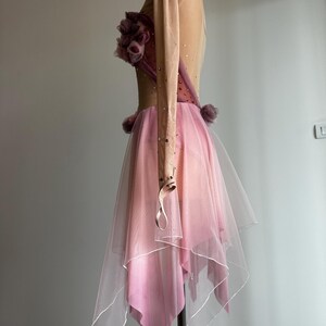 May include: A long-sleeved dance dress featuring a nude-colored bodice with scattered rhinestones, a cluster of mauve fabric flowers at the chest, and a layered, asymmetrical pink tulle skirt with a white hemline, displayed on a mannequin.