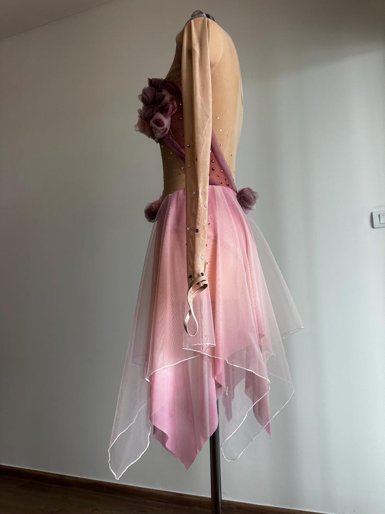 May include: A long-sleeved dance dress featuring a nude-colored bodice with scattered rhinestones, a cluster of mauve fabric flowers at the chest, and a layered, asymmetrical pink tulle skirt with a white hemline, displayed on a mannequin.