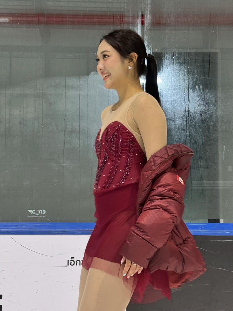 May include: A maroon figure skating dress featuring a sheer nude-colored neckline and bodice overlay, adorned with intricate dark red beadwork and sequins. The skirt is made of layered maroon fabric, paired with a matching maroon quilted puffer jacket.