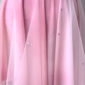 May include: A pink dress featuring layers of sheer fabric, two fabric rosettes at the waist, and scattered small silver embellishments. The skirt has a flowing, layered design with a white hemline detail, displayed on a dress form mannequin.