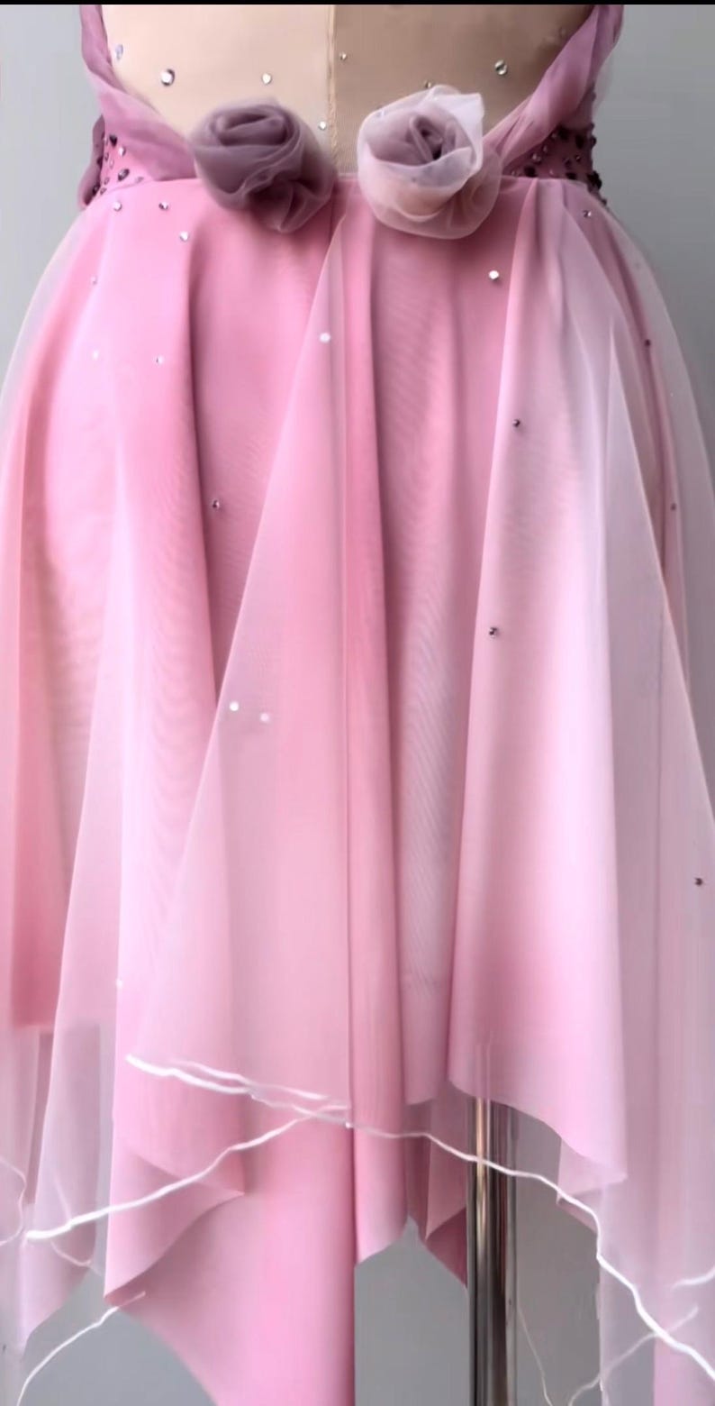 May include: A pink dress featuring layers of sheer fabric, two fabric rosettes at the waist, and scattered small silver embellishments. The skirt has a flowing, layered design with a white hemline detail, displayed on a dress form mannequin.