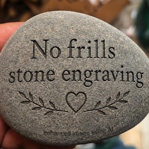 Puede incluir: Una piedra de río lisa, gris y de forma ovalada con el texto grabado "No frills stone engraving" en una fuente serif. Debajo del texto, hay un símbolo de corazón grabado flanqueado por dos ramas con hojas, con el texto pequeño "enhanced image using AI" en la parte inferior.