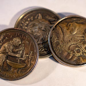 May include: Three circular bronze-toned metal medallions featuring embossed relief designs. One depicts a person working at a workbench, another shows a detailed animal head, and the third displays a complex scene with mechanical gears and figures.