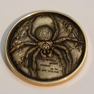 Puede incluir: Una moneda redonda de color bronce con una imagen detallada en tres dimensiones de una araña. La araña está mirando hacia adelante con las patas extendidas. La moneda tiene una inscripción que dice "Trey Wingy" y otro texto.