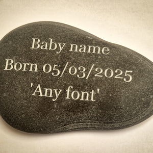 May include: Dark grey engraved stone with the text "Baby name Born 05/03/2025 'Any font'". The stone has a smooth, rounded shape, likely a memorial or keepsake.