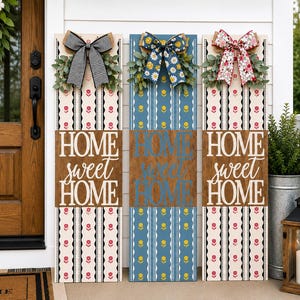 May include: Three wooden HOME SWEET HOME signs with decorative bows and floral accents. The signs feature patterned backgrounds in cream and blue, with the words "HOME SWEET HOME" in a script font.