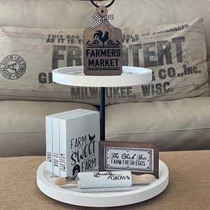May include: A white tiered tray with a black metal stand. The top tier has a wooden sign that says "Farmers Market" with a black rooster and a red and white checkered bow. The middle tier has three white signs that say "Farm Sweet Farm", "Welcome to our Farm", and "Farm". The bottom tier has a wooden sign that says "The Chick Inn Farm Fresh Eggs" and a rolling pin that says "Locally Grown".