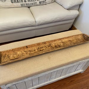 May include: A long, rustic wooden dough bowl with a natural finish. The bowl is rectangular with rounded ends and sits on a beige upholstered bench. The bench has a white, distressed wooden base. The bowl is approximately 36 inches long.