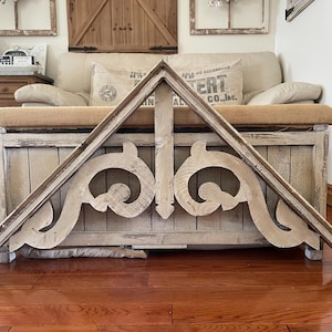 May include: A distressed, wooden architectural bracket in a triangular shape. The bracket features ornate, scroll-like carvings and a weathered, off-white finish. It is resting on a wooden chest with a neutral-toned cushion.