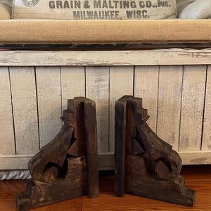 Peut inclure: Une paire de consoles en bois brun foncé présentant un design architectural à gradins avec des détails courbes en forme de volute. Ces supports structurels ont une finition vieillie et patinée avec un grain de bois visible et des sculptures.