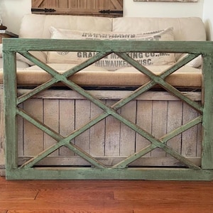 May include: A distressed wooden frame with a crisscross pattern, painted in a faded green colour. The frame is positioned in front of a wooden chest and a beige sofa, creating a rustic, farmhouse aesthetic.