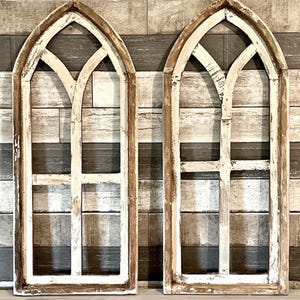 May include: Two decorative wooden window frames with an arched top. The frames are painted white with a distressed finish, and the trim is a natural wood colour. The windows are set against a backdrop of wooden planks.