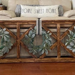 May include: A wooden frame with a diamond lattice design, featuring three green wreaths with black and white ribbon accents. The frame is mounted on a wall above a sofa with a "Home Sweet Home" cushion.