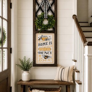 May include: A vertical rectangular wall sign with a dark wood frame, featuring a black and white buffalo check bow at the top and a green wreath. The sign displays the text "HOME IS where my HONEY Bee" with illustrations of a honey jar and a bee.