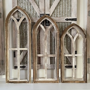 May include: Three rustic wooden arched window frames with a distressed white paint finish. The frames are arranged in a row, with the largest frame on the left and the smallest frame on the right.