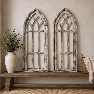 May include: Two tall, arched window frames featuring a distressed white and black finish. Each frame has a pointed top and a grid of vertical and horizontal muntins, creating a multi-pane design. The frames are positioned upright side by side.