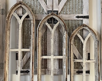 May include: Three distressed, arched wooden window frames in varying sizes, with a weathered white and brown finish. The frames are set against a backdrop of a rustic, barn-style door with metal accents. The overall aesthetic is farmhouse style.