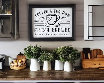 May include: A black and white sign with the text "COFFEE & TEA BAR" and "OPEN 24/7" and "SELF SERVE" and "FRESH BREWED" and "love brews here". The sign is framed in black and has a picture of a coffee cup with a heart on it.