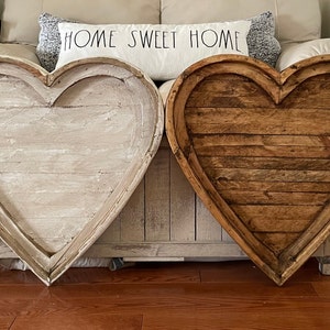 May include: Two large wooden heart-shaped signs, one painted white and the other stained brown, are displayed on a couch. The signs are made of wood planks and have a rustic, farmhouse style.