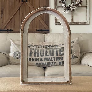 May include: A distressed wooden arched window frame, painted in white and brown. The frame is empty, with a vintage-style pillow visible through the opening. The pillow has text that reads "FROEDTE GRAIN & MALTING MILWAUKEE, WIS".