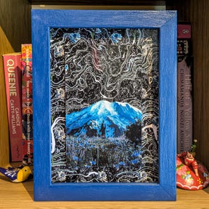 Puede incluir: Una impresión artística enmarcada que presenta un paisaje montañoso en tonos azules y blancos. La obra de arte está enmarcada en un marco de madera azul. El fondo es negro con patrones blancos en espiral. Libros y objetos decorativos son visibles.