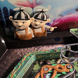 May include: Two small figurines of boys wearing striped hats, striped shirts, and brown overalls. One figure points to the right. The figurines are on a pinball machine with a colorful, whimsical design featuring mushrooms and other fantasy elements.