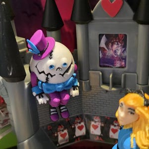 May include: A whimsical Alice in Wonderland-themed playset featuring a Humpty Dumpty figure with a purple top hat, a castle with a heart, and an Alice figure in a blue dress. The scene is set with playing card soldiers.