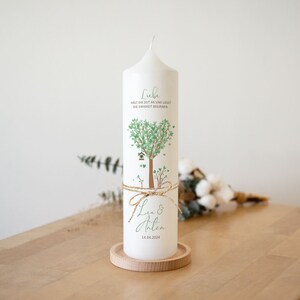 Könnte beinhalten: Weiße Stumpenkerze mit einem grünen herzförmigen Baumdesign und dem Text "Liebe hält die Zeit an und lässt die Ewigkeit beginnen." Die Kerze ist mit einer Schnur gebunden und trägt die Namen "Lea & Anton" sowie das Datum "14.04.2024" aufgedruckt.