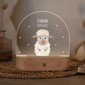 Könnte beinhalten: Ein weißes Acryl-Nachtlicht mit einem Holzsockel. Das Licht zeigt ein Cartoon-Schaf und den Text "FABIAN 23.04.2023".