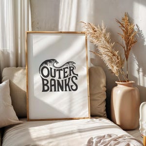 May include: A black and white print with the text "Outer Banks" in a stylized font, with waves behind the text. The print is framed in a light wood frame and is hanging on a wall.