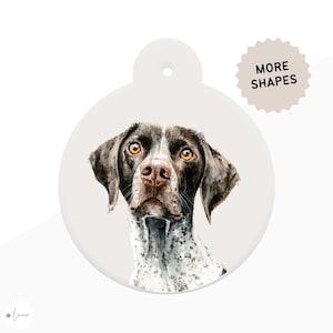 May include: A round, off-white pet tag featuring a watercolor illustration of a dog's face. The dog has brown and white fur, with amber eyes and a brown nose. The tag has a hole at the top for attachment and the text "MORE SHAPES".