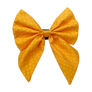 May include: A large yellow bow with a polka dot pattern. The bow is made of fabric and has a black elastic band in the centre.