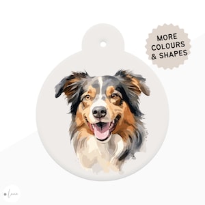 Puede incluir: Una etiqueta redonda y blanquecina para mascotas con un retrato de estilo acuarela de un perro pastor australiano. El perro tiene pelaje marrón, negro y blanco, con una expresión feliz. El texto en la etiqueta dice "MORE COLOURS & SHAPES".