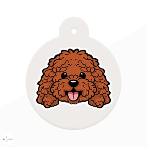 May include: A circular, pale grey pet tag with a cartoon depiction of a brown dog's face. The dog has a curly coat, black eyes, a black nose, and a pink tongue. The tag has a hole at the top for attachment.