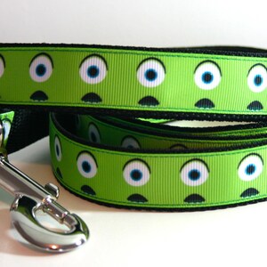 May include: Green dog lead with a black border and a repeating pattern of cartoon eyes. The lead has a silver clip.