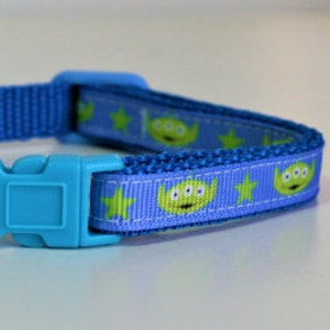 May include: A blue and purple dog collar with a repeating pattern of green alien faces and yellow stars. The collar has a blue plastic buckle and a silver D-ring.