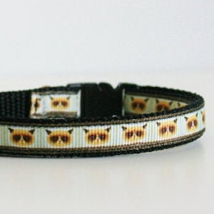 May include: A black cat collar with a white background and a repeating pattern of brown and white cat faces. The collar has a black buckle.