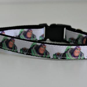 Puede incluir: Un collar para perros con un patrón repetido de un personaje de dibujos animados en verde, morado y naranja sobre un fondo blanco. El collar tiene una hebilla negra y un interior negro.