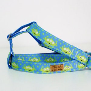 May include: A blue and green dog harness with a repeating pattern of three-eyed aliens. The harness has a silver buckle and two silver D-rings. The text "Bohe's Collars" is printed on a brown tag attached to the harness.