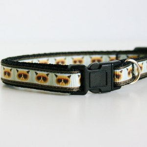 May include: A black cat collar with a white background and a repeating pattern of grumpy cat faces. The collar has a black plastic buckle and a silver metal ring.