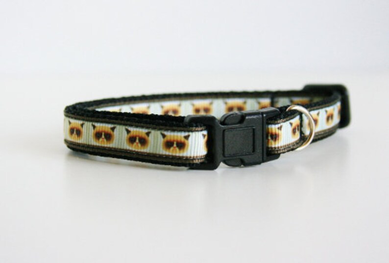 May include: A black cat collar with a white background and a repeating pattern of grumpy cat faces. The collar has a black plastic buckle and a silver metal ring.