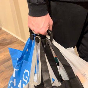 Puede incluir: Una persona sostiene un porta bolsas de plástico negro con varias bolsas de la compra sujetas. Las bolsas son una bolsa reutilizable azul, una bolsa de plástico blanca y una bolsa reutilizable negra.