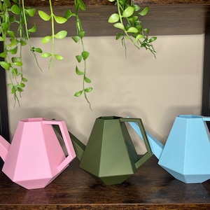 Puede incluir: Tres regaderas geométricas en rosa, verde y azul. Las regaderas son de plástico y tienen un pico y un asa.