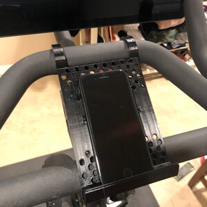 May include: Black phone holder mounted on the handlebars of a stationary exercise bike. The holder is made of black plastic and has a grid pattern. The phone is in a vertical position.